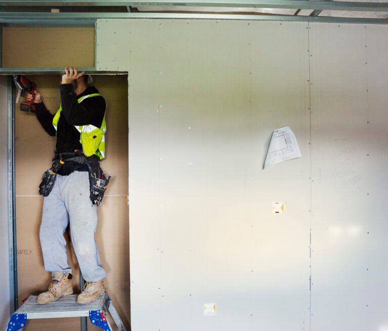 A carpenter in work clothes on a construction site, using an electric drill.