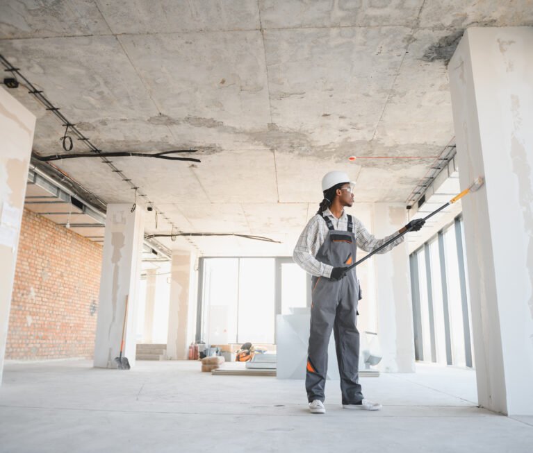Construction worker painting walls and columns in a building under construction, using long brush and wearing safety equipment