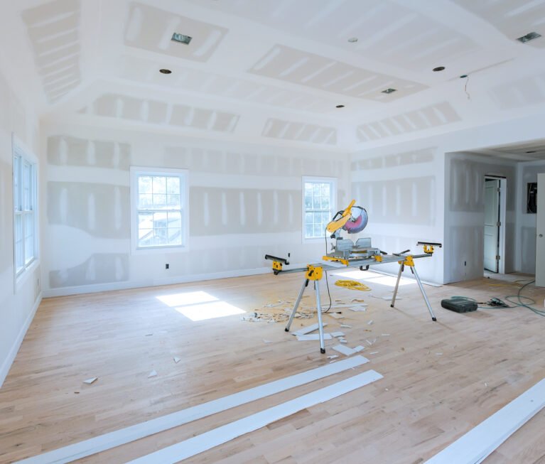 Spacious, unfinished room with drywall partially installed miter saw set up in center on stand, surrounded by wood trim pieces construction debris at hardwood floor.
