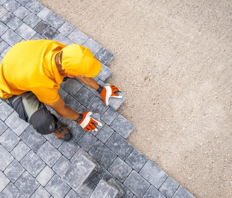 A construction worker is carefully placing gray paving stones onto a prepared surface, ensuring alignment and stability. Dressed in an orange shirt and gloves, he demonstrates skilled craftsmanship in a sunny outdoor environment.